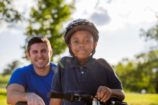 Big standing behind Little on a bike with helmet on